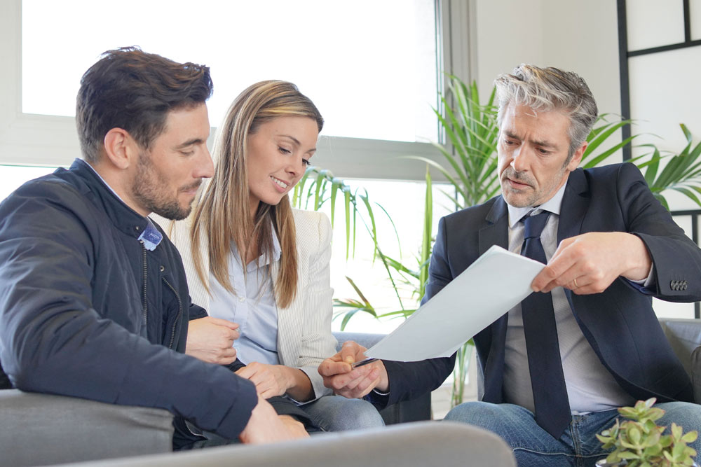 Drei Personen sitzen zusammen, eine Frau und ein Mann schauen auf ein Dokument, das ein Mann in Anzug hält. Pflanzen sind im Hintergrund zu sehen.